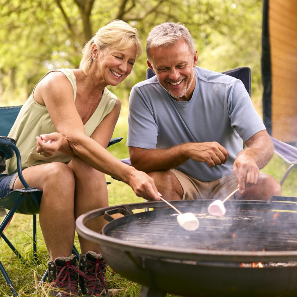 Senior Couple Camping In Countryside With RV Toasting Marshmallows Outdoors On Fire