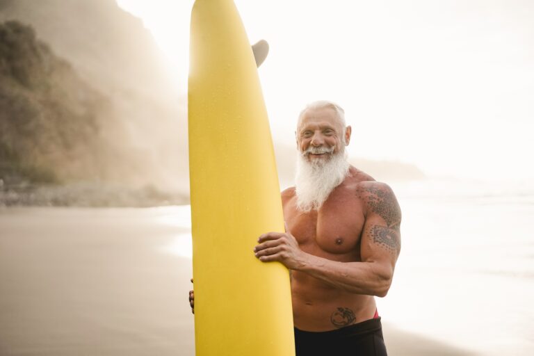 Senior surfer man holding surf board on the beach at sunset - Focus on face